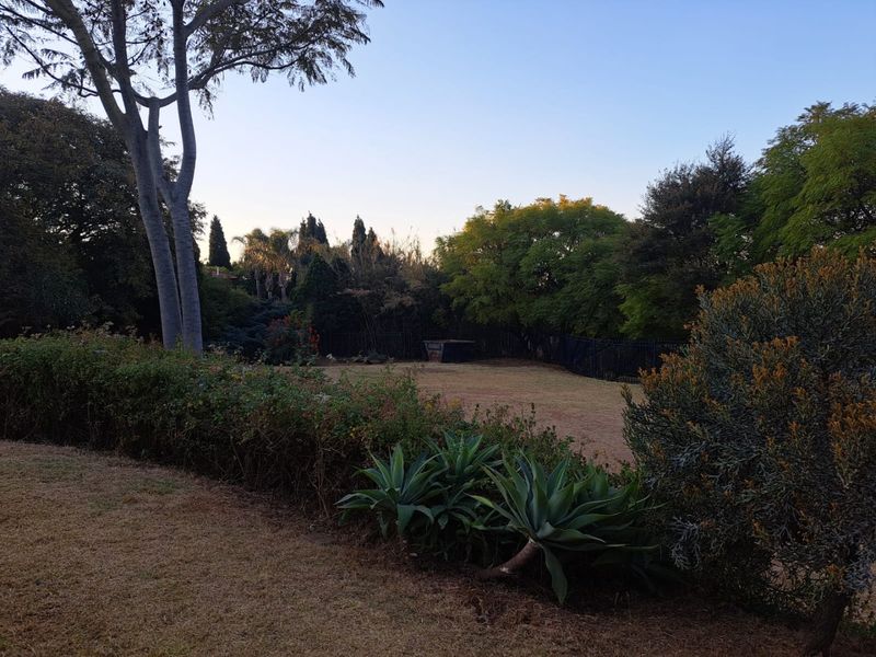 A spacious residential garden featuring mature trees, established hedging, and a well-defined lawn area enclosed by perimeter fencing.
