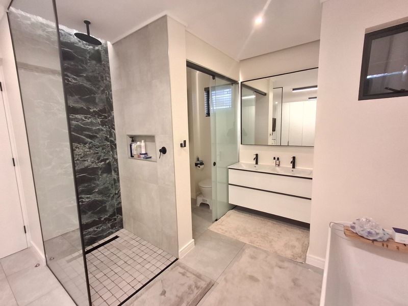 Modern bathroom featuring a walk-in shower with marble-look tiling, a double vanity with black fixtures, and a separate enclosed toilet area.