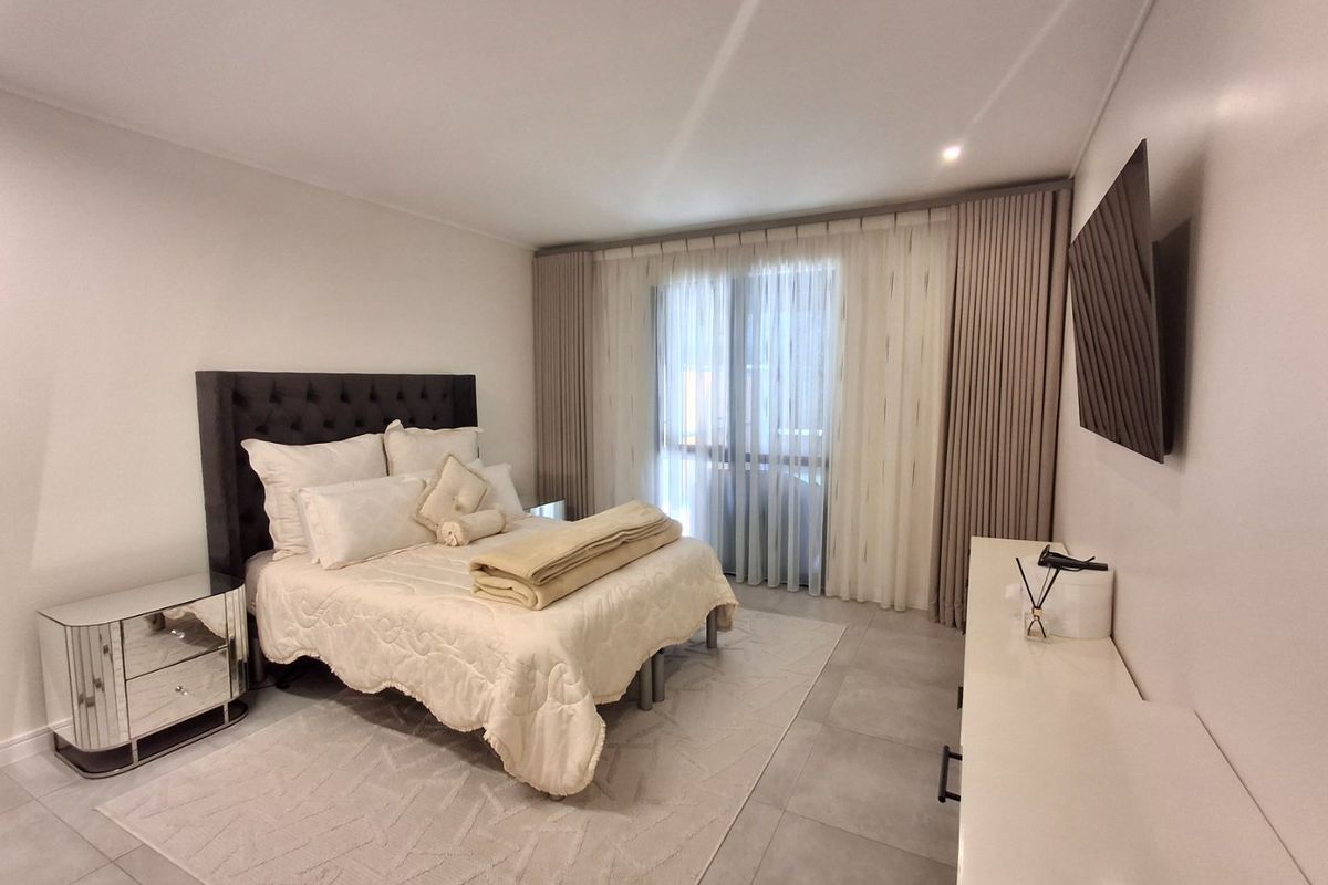 A well-maintained bedroom featuring neutral tiled flooring, recessed lighting, and large sliding glass doors with floor-to-ceiling curtains providing access to the exterior.
