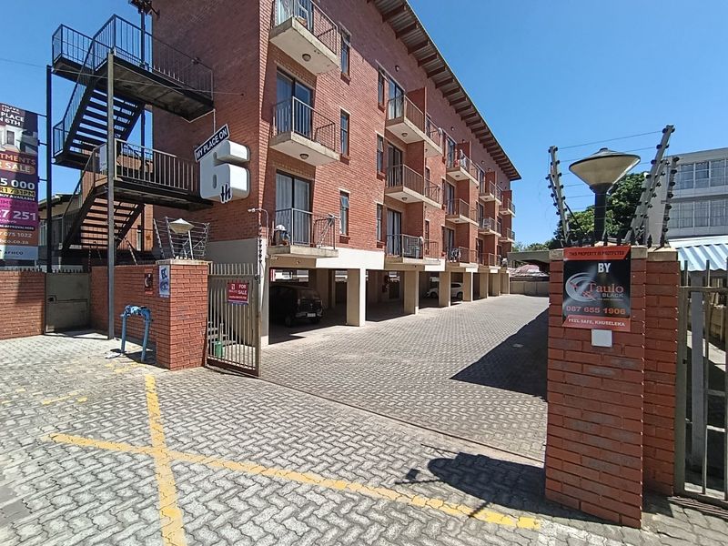 Exterior view of a multi-story brick apartment building featuring balconies, a paved driveway, covered parking, and perimeter security fencing.