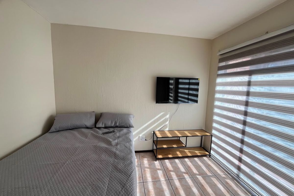 A bedroom featuring tiled flooring, wall-mounted television, and modern roller blinds, providing a functional and well-lit living space.