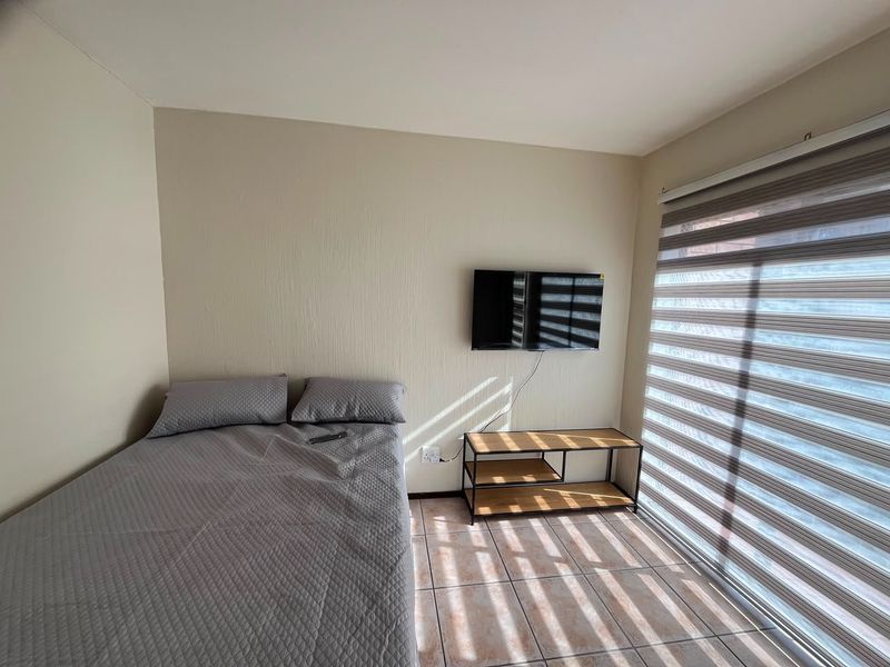 A bedroom featuring tiled flooring, wall-mounted television, and modern roller blinds, providing a functional and well-lit living space.