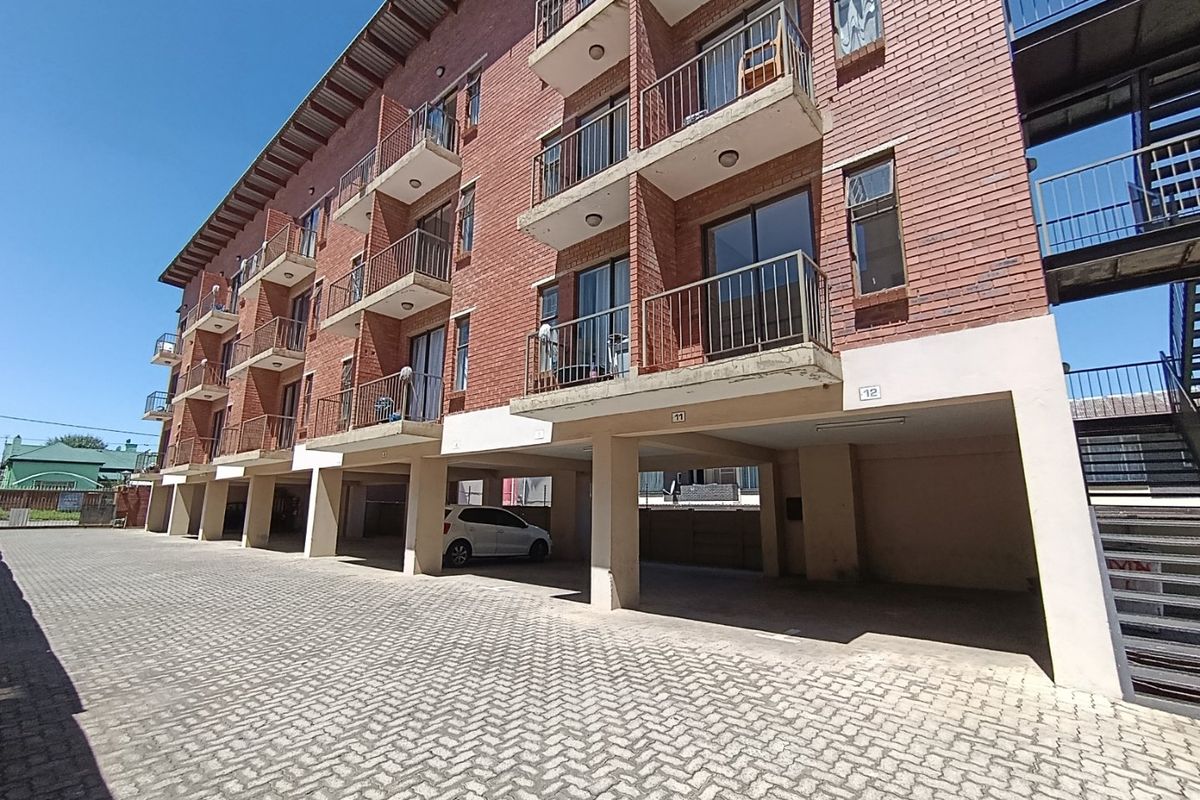 Exterior view of a multi-story brick apartment building featuring private balconies and ground-level covered parking bays with paved driveway access.