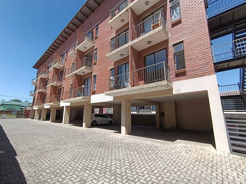 Exterior view of a multi-story brick apartment building featuring private balconies and ground-level covered parking bays with paved driveway access.