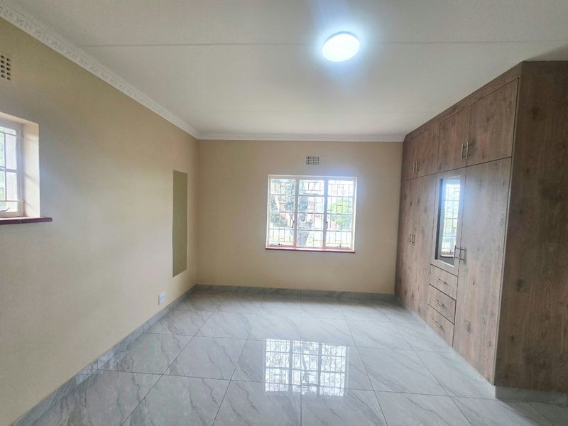 A bright, empty bedroom featuring modern tiled flooring, neutral walls, and extensive built-in wooden cupboards with integrated storage.