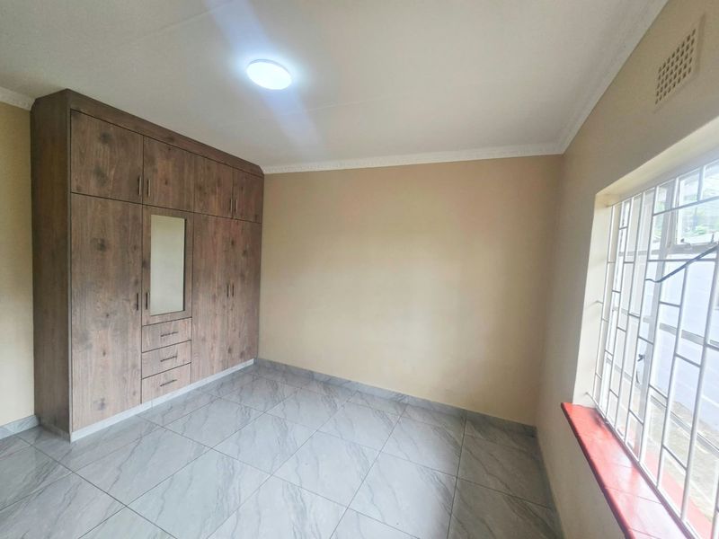 A clean, empty bedroom featuring modern wood-look built-in cupboards, neutral wall paint, and light grey tiled flooring with security bars on the window.
