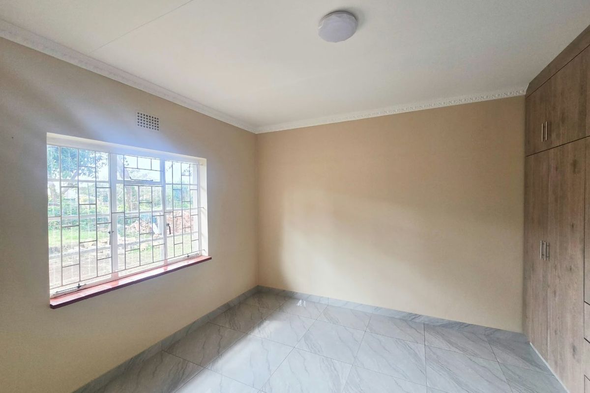A bright, empty bedroom featuring modern tiled flooring, built-in wooden cupboards, and a window fitted with security burglar bars.