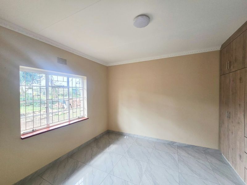 A bright, empty bedroom featuring modern tiled flooring, built-in wooden cupboards, and a window fitted with security burglar bars.
