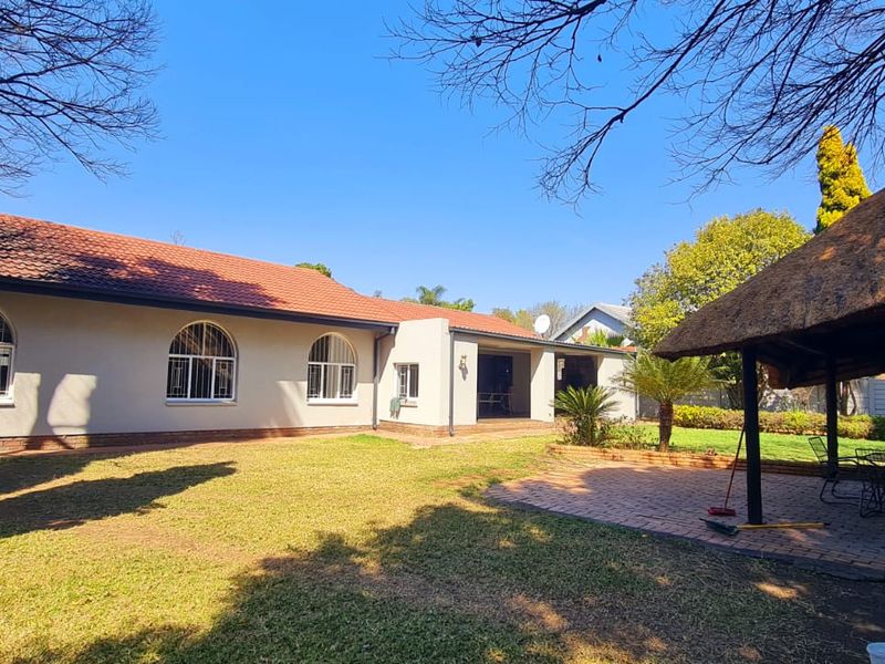 Spacious rear garden featuring a paved patio area, a traditional thatched lapa, and a well-maintained lawn with established trees.