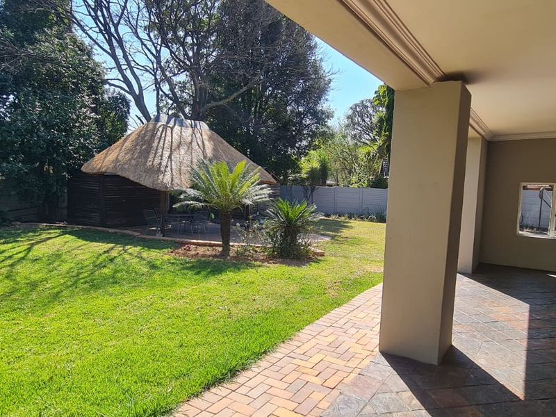 A covered patio overlooking a spacious garden featuring a thatched lapa and paved pathways, offering a pleasant outdoor entertainment space.