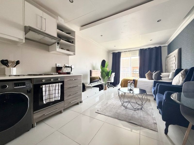 Modern open-plan studio apartment featuring a compact kitchen with built-in appliances, tiled flooring, and a bright living and sleeping area with balcony access.