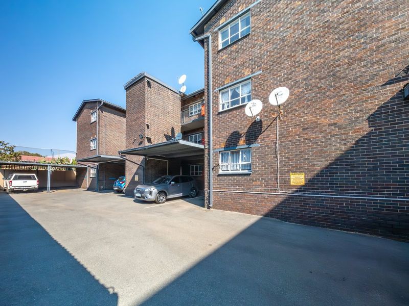 Exterior view of a multi-story brick apartment building featuring covered parking bays and an asphalt driveway.