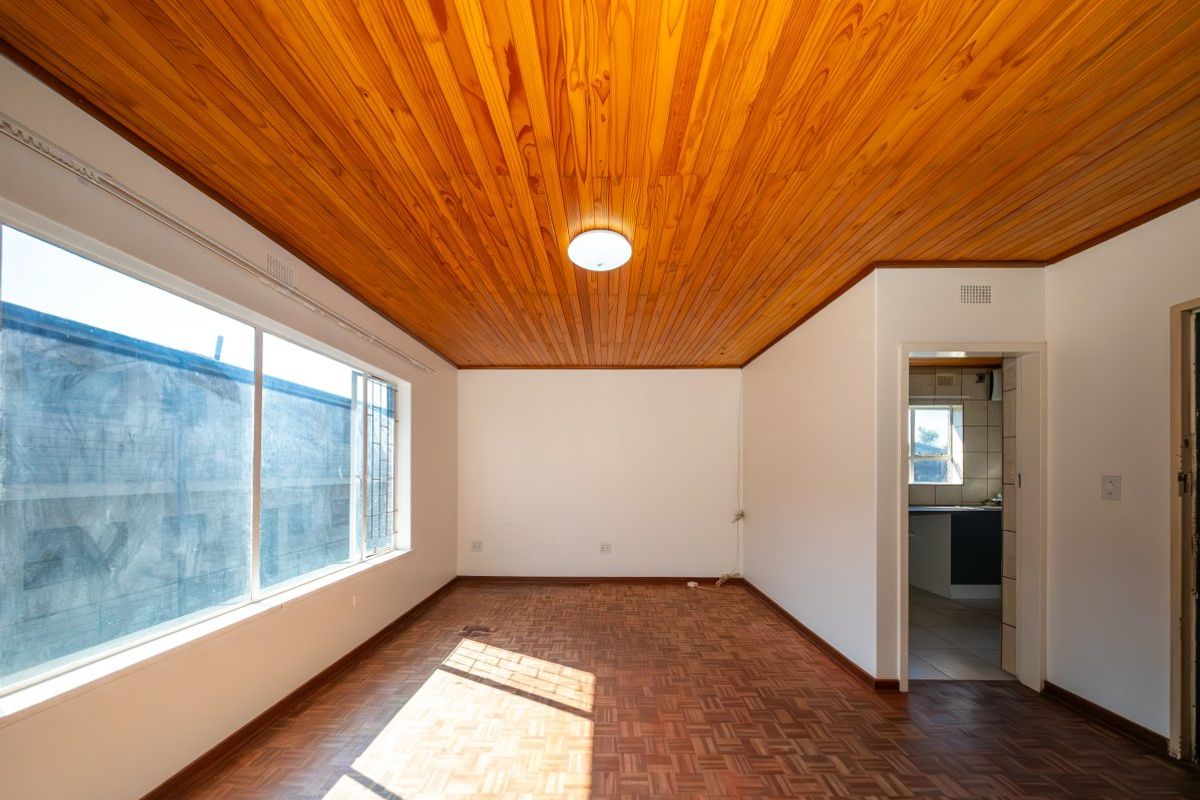 An empty living room featuring classic parquet flooring and a warm wooden tongue and groove ceiling, with natural light from a large window.