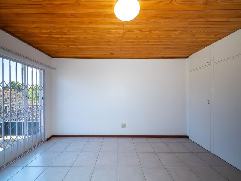An empty bedroom featuring a timber ceiling, tiled flooring, and built-in cupboards, with access to a balcony through a secure sliding door.