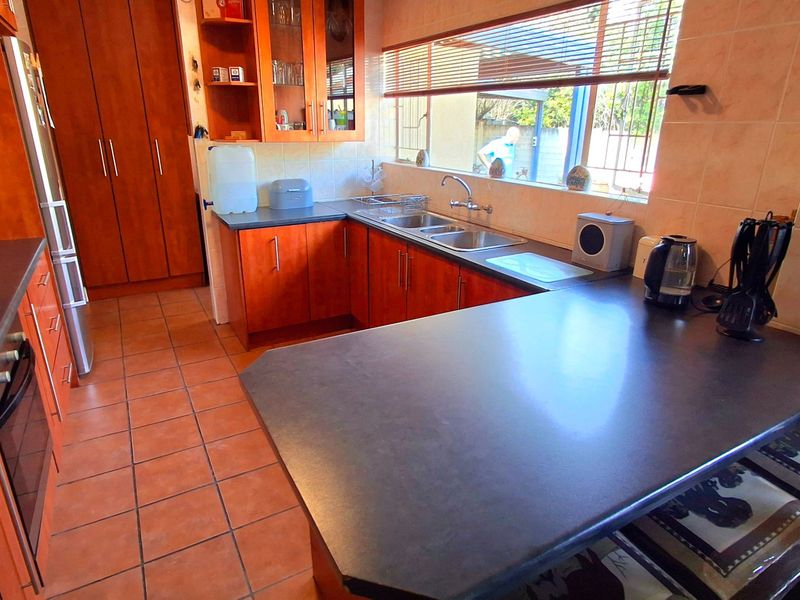 A functional kitchen featuring warm-toned wooden cabinetry, dark laminate countertops, a double basin sink, and tiled flooring with a practical layout.