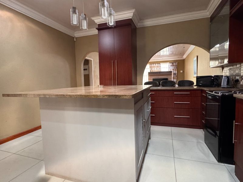 A spacious kitchen featuring a central island with stone countertops, dark wood cabinetry, tiled flooring, and an open archway leading to the living area.