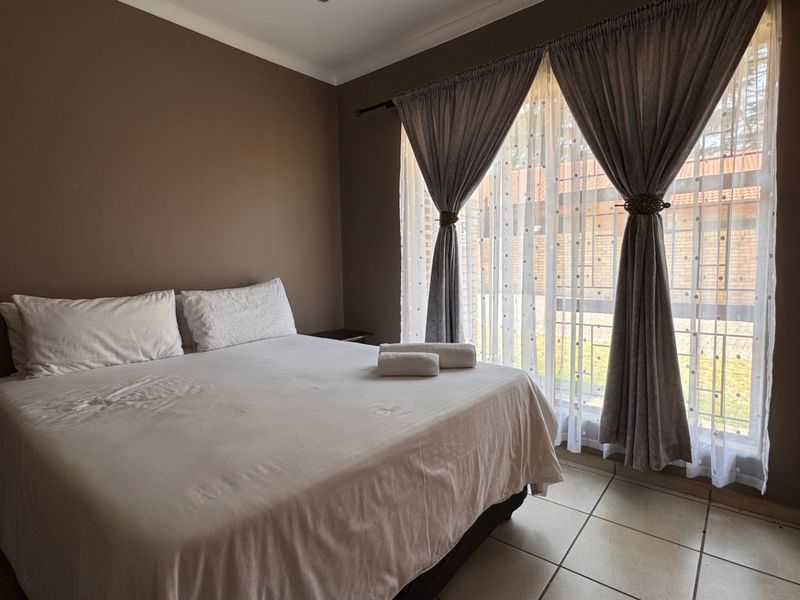 A bedroom featuring tiled flooring, neutral wall tones, and a built-in wardrobe, with natural light provided by a large window with curtain treatments.