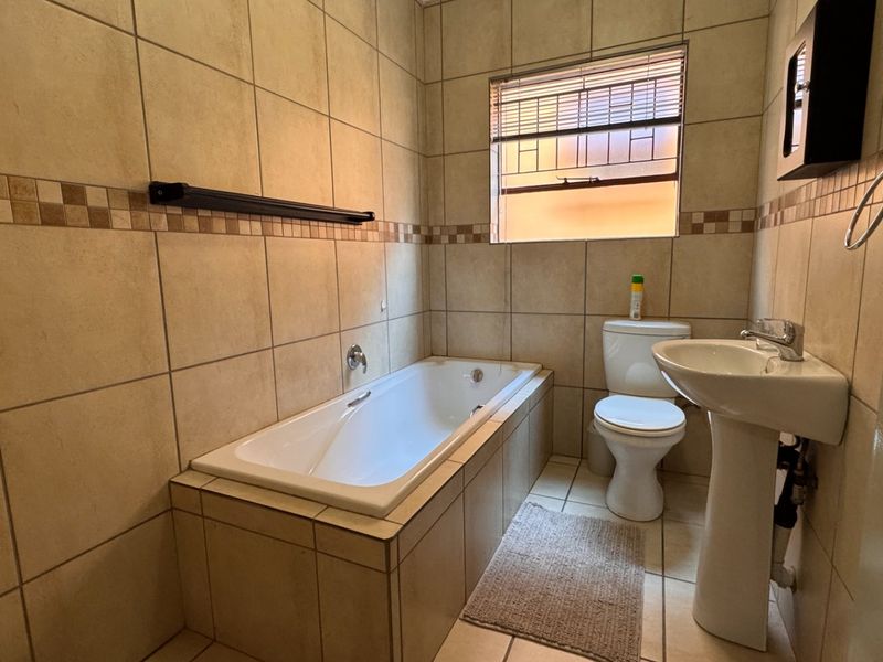 A functional bathroom featuring a built-in bathtub, pedestal basin, and toilet, finished with neutral wall and floor tiling.