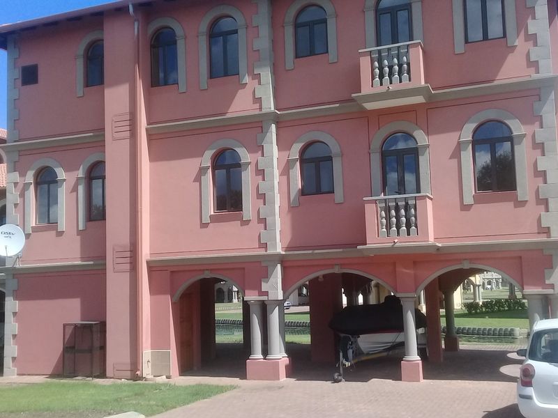 A multi-story residential building featuring a distinctive pink facade, arched windows, decorative balustrades, and a covered ground-level parking area with arched columns.