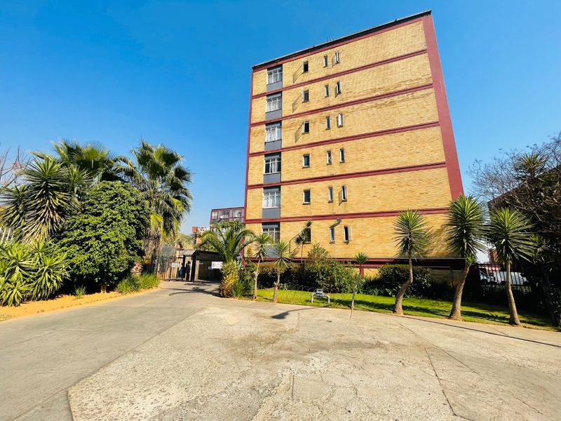 Exterior view of a multi-story brick apartment building featuring a paved driveway, landscaped garden area, and secure gated entrance.