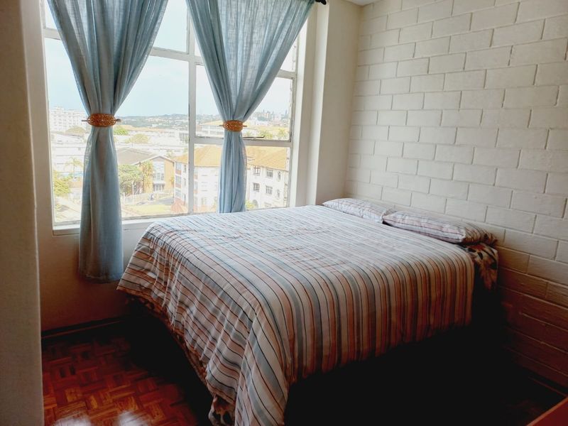 A bedroom featuring parquet flooring, an exposed brick feature wall, and a large window offering an elevated urban view.