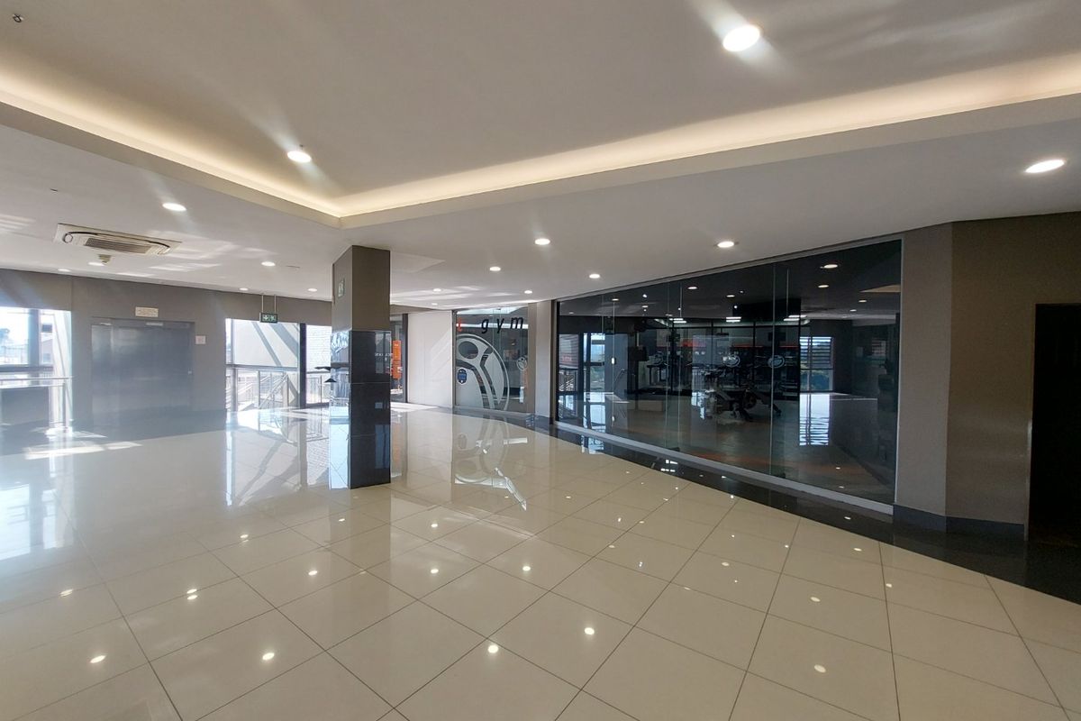 Spacious commercial hallway featuring polished tiled flooring, recessed and cove lighting, and a glass-partitioned entrance to a gym facility.