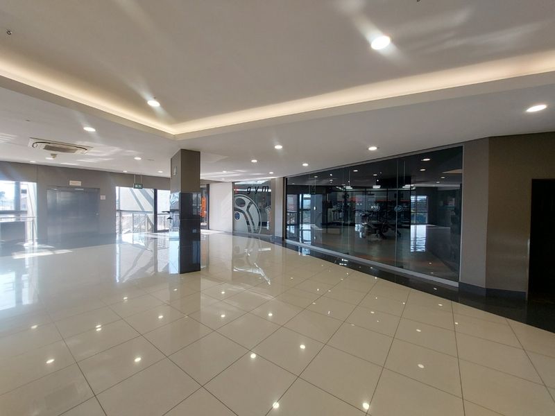 Spacious commercial hallway featuring polished tiled flooring, recessed and cove lighting, and a glass-partitioned entrance to a gym facility.