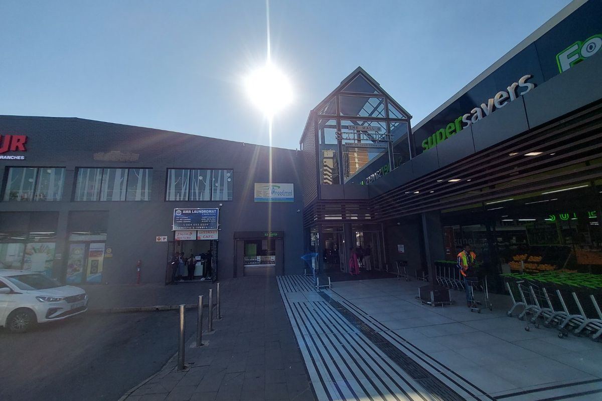 Front exterior view of a commercial shopping center featuring a glass atrium entrance, paved pedestrian walkway, and prominent retail signage.