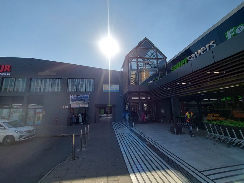 Front exterior view of a commercial shopping center featuring a glass atrium entrance, paved pedestrian walkway, and prominent retail signage.