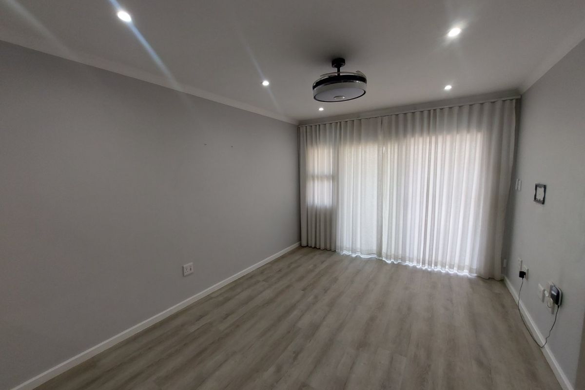 A bright, empty living room featuring modern laminate flooring, recessed downlights, a ceiling fan, and floor-to-ceiling curtain tracks.