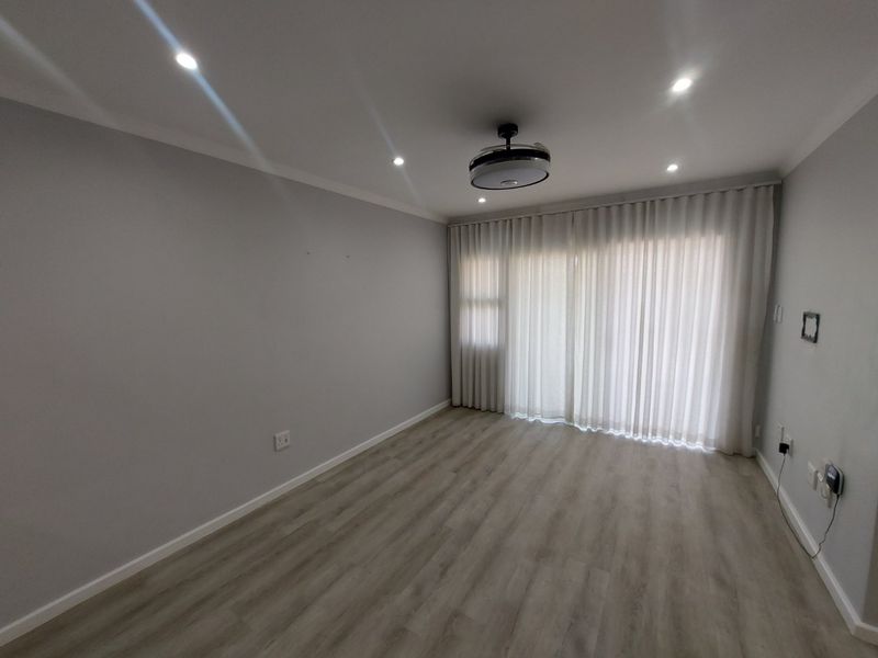 A bright, empty living room featuring modern laminate flooring, recessed downlights, a ceiling fan, and floor-to-ceiling curtain tracks.
