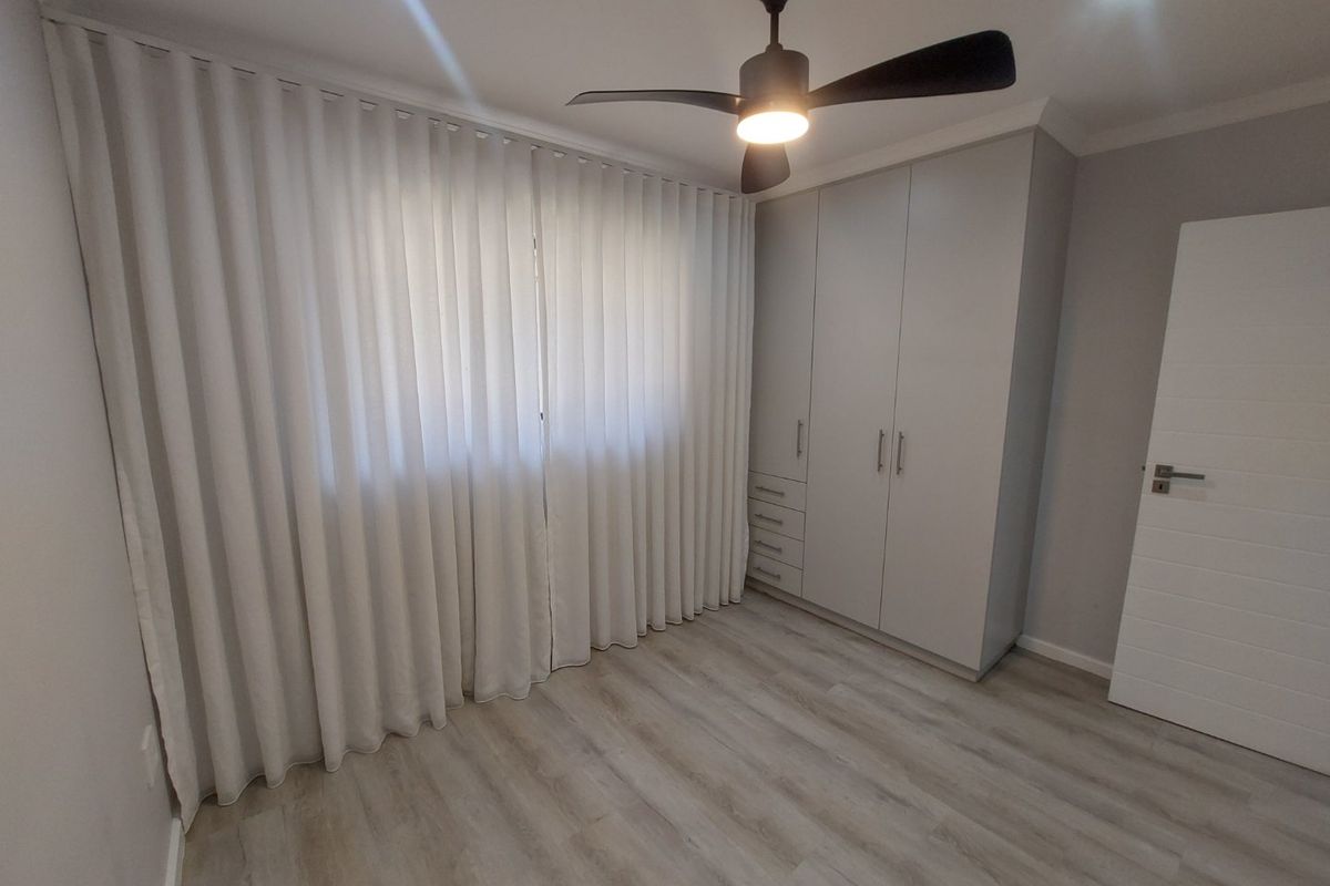 A clean, modern bedroom featuring light laminate flooring, built-in cupboards, a ceiling fan, and recessed lighting with floor-to-ceiling curtains.