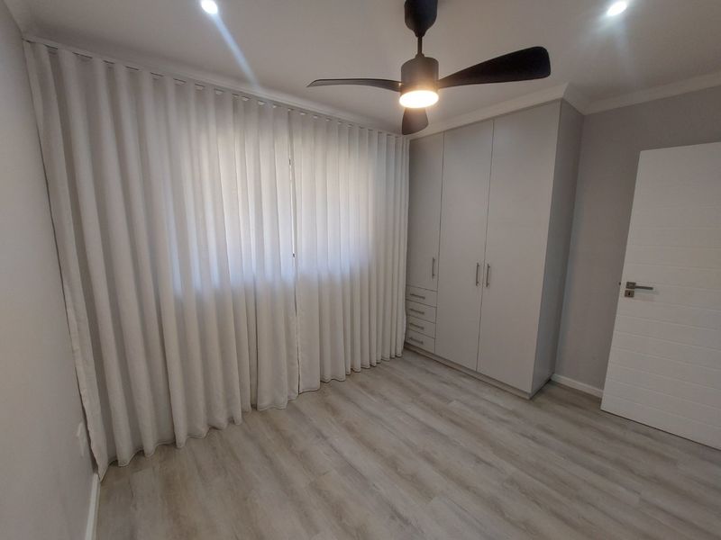 A clean, modern bedroom featuring light laminate flooring, built-in cupboards, a ceiling fan, and recessed lighting with floor-to-ceiling curtains.
