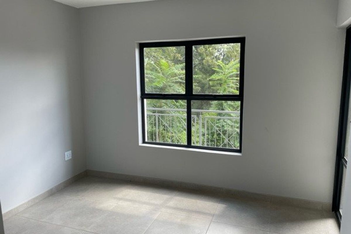 A bright, empty bedroom featuring neutral tiled flooring, clean grey walls, and a large window overlooking greenery.