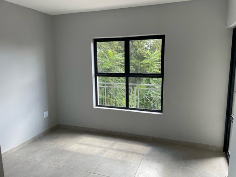 A bright, empty bedroom featuring neutral tiled flooring, clean grey walls, and a large window overlooking greenery.