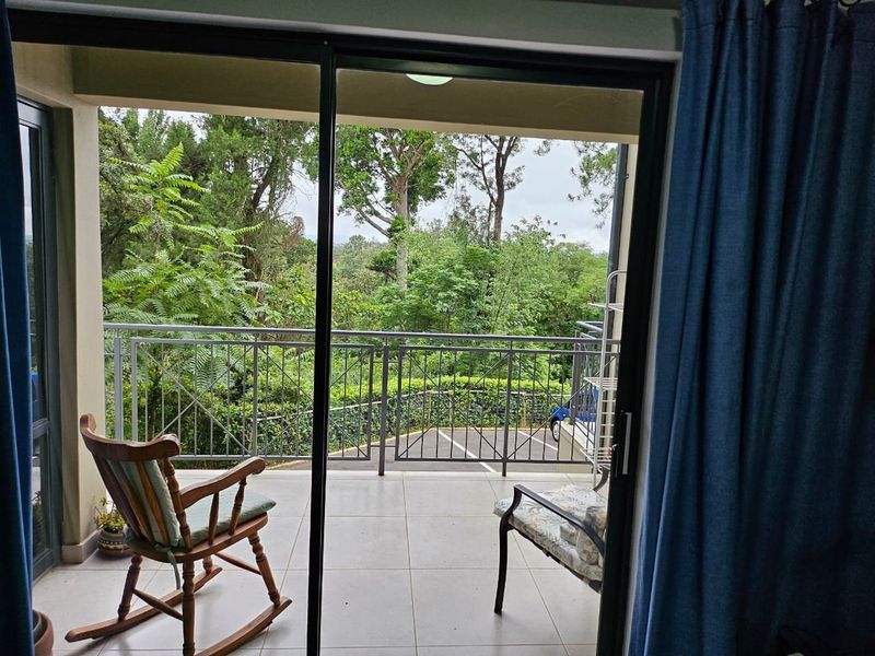 A covered balcony featuring tiled flooring and a metal balustrade, offering a pleasant view of the surrounding greenery and trees.