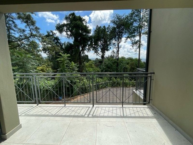 A spacious covered balcony featuring tiled flooring and a secure metal balustrade, offering pleasant views of the surrounding trees and landscape.