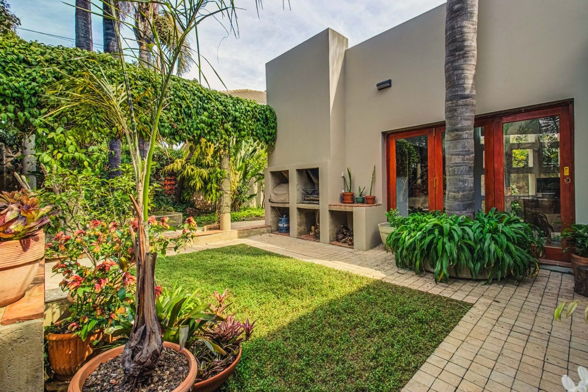 A well-maintained rear garden featuring a paved patio area, a built-in braai, and established greenery with a wooden-framed entrance.