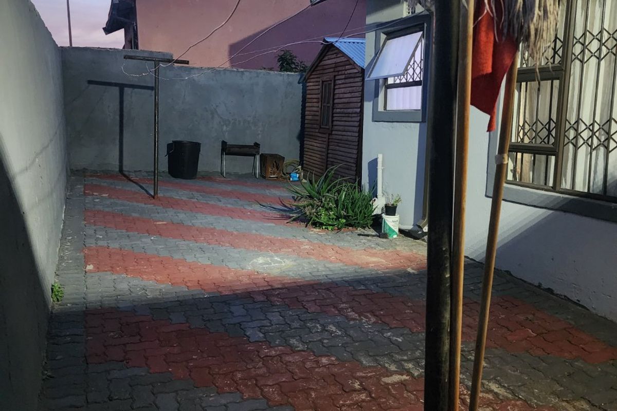 A paved rear exterior area featuring boundary walls, security bars on windows, and a small storage shed, showing signs of wear.