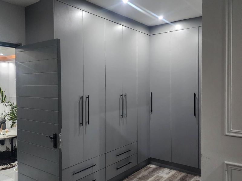 A modern bedroom featuring extensive grey built-in cupboards, laminate wood-look flooring, and recessed ceiling lighting with decorative cornice detailing.