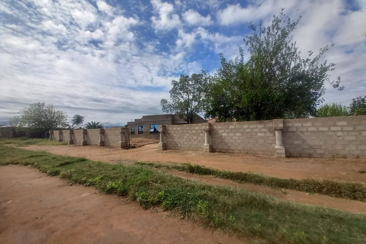 Street view of the property showing an unfinished perimeter wall with concrete pillars and unpaved road access.