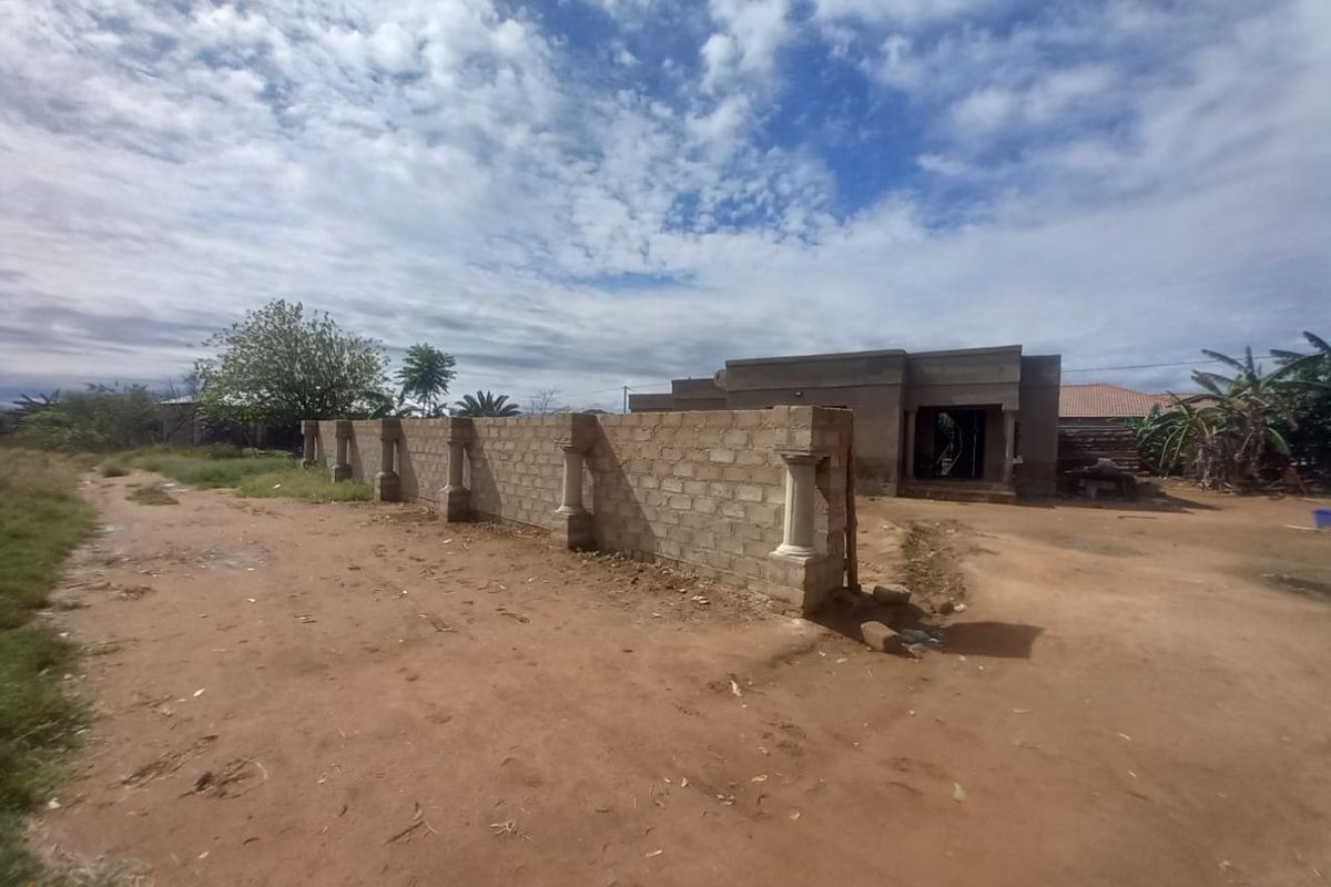 Front view of a property under construction featuring an unfinished concrete block boundary wall with decorative pillars and an unpaved driveway.