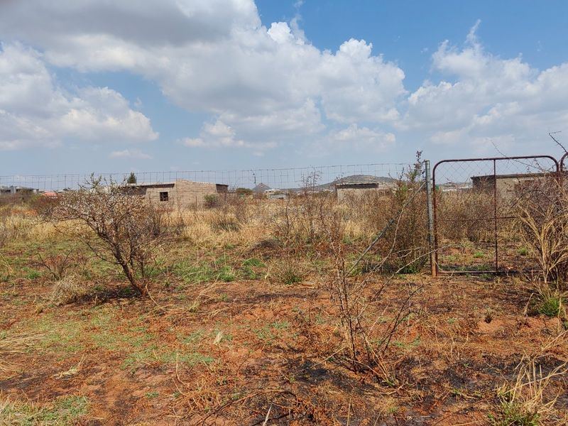 A view of a vacant residential plot featuring wire mesh fencing and a metal gate, with dry vegetation and surrounding structures in the distance.