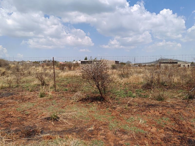 A view of a vacant residential plot featuring dry natural vegetation and perimeter wire mesh fencing.
