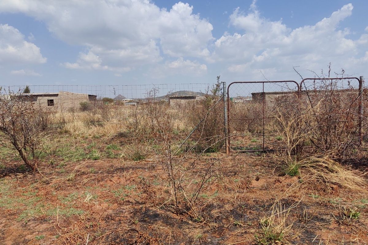 A view of a vacant residential plot featuring wire mesh fencing and a metal gate, with dry, overgrown vegetation and scorched ground.
