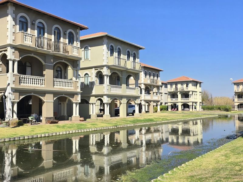 A scenic view of multi-story residential villas featuring classic architectural details, balconies, and arched windows, situated along a tranquil canal with manicured lawns.