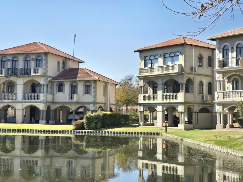 A view of multi-story residential units featuring Mediterranean-style architecture, terracotta roofs, and balconies overlooking a tranquil pond and landscaped grounds.