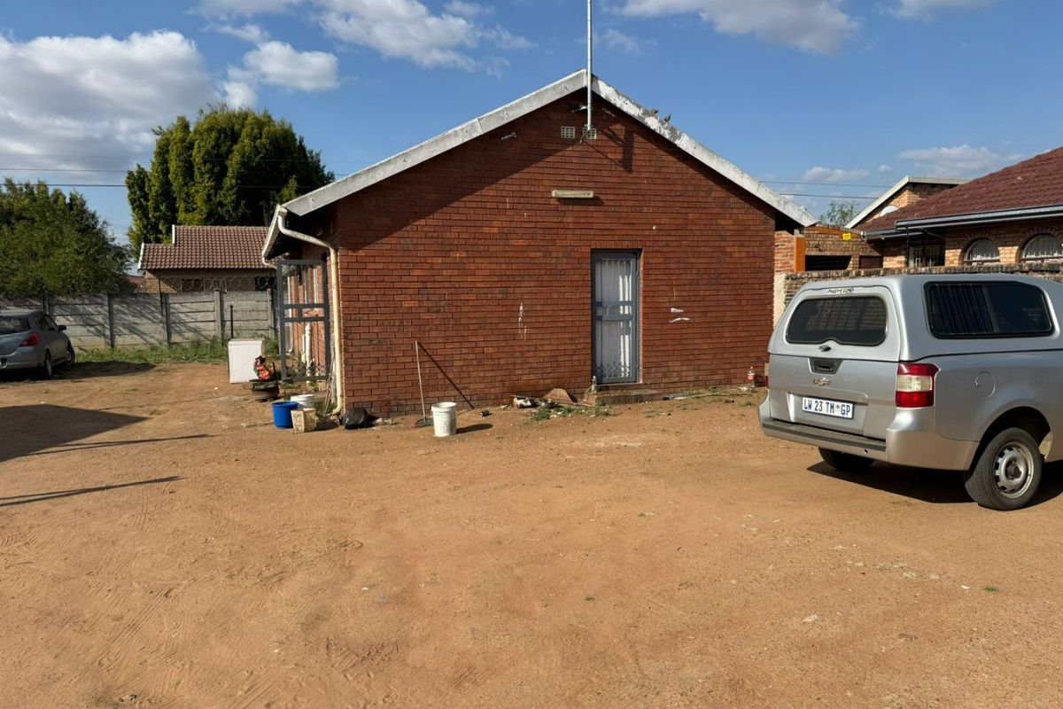 Rear view of a brick house featuring a gabled roof and security gate, set within an unpaved yard with a perimeter boundary wall.