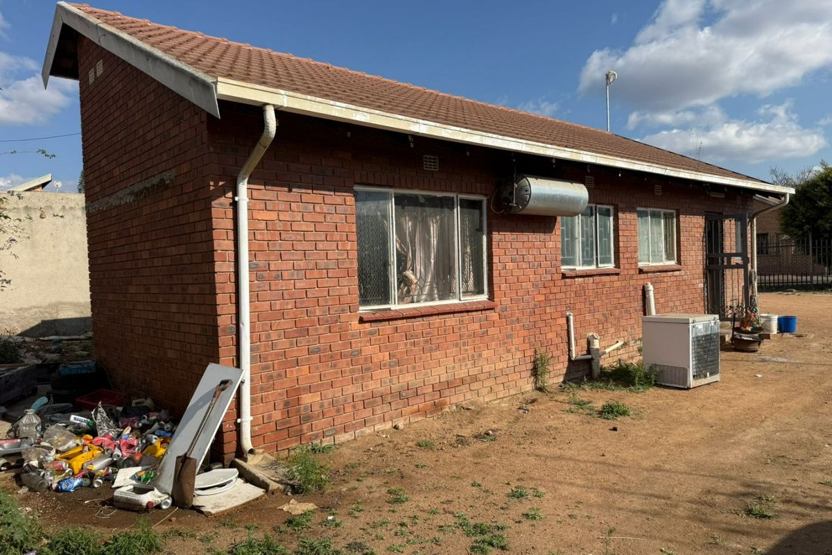 Rear view of a face brick house featuring a tiled roof, external geyser, and security gate, situated on an unmaintained plot.