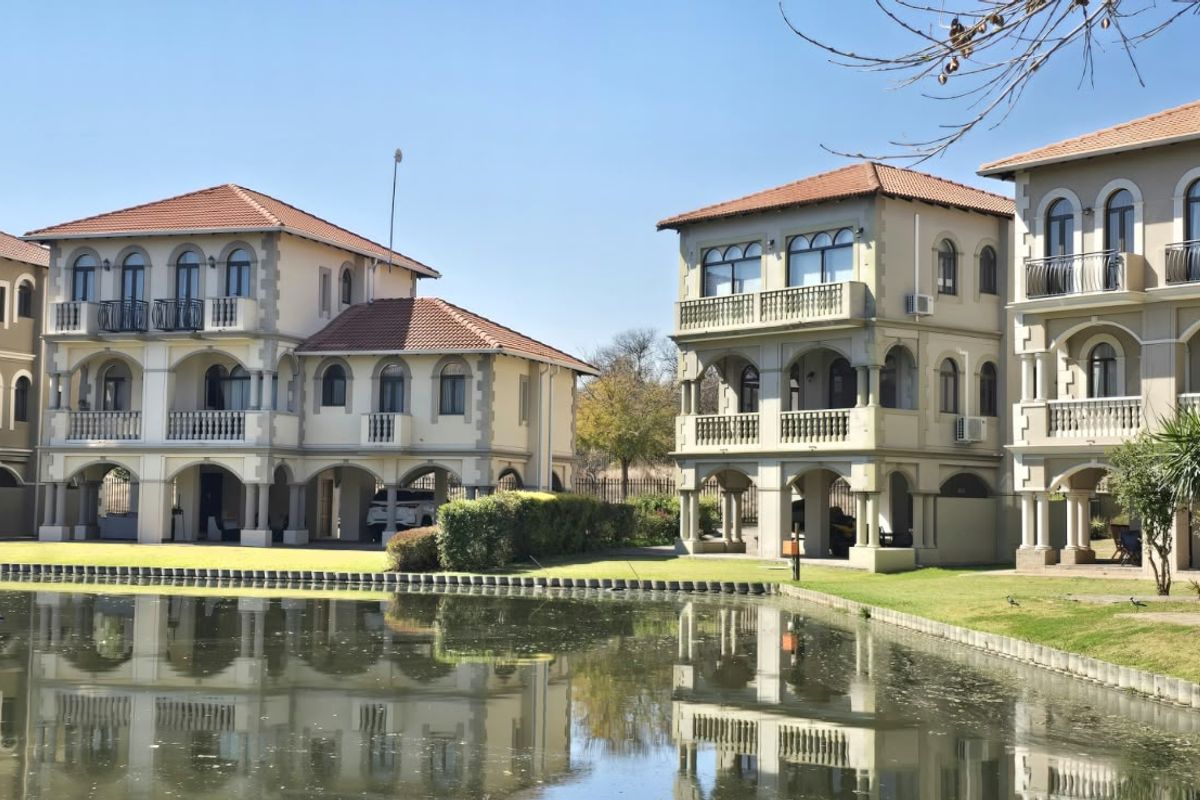 A view of multi-story residential units featuring Mediterranean-style architecture, terracotta roofs, and balconies overlooking a tranquil pond and landscaped grounds.